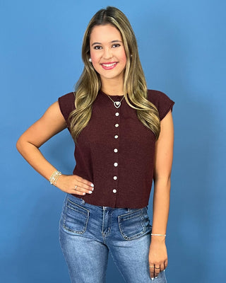 A woman with blonde hair wearing a sleeveless maroon button-up knit top and blue jeans, standing against a solid blue background.