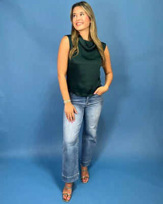 A woman modeling a dark green sleeveless cowl-neck top and light-wash blue jeans