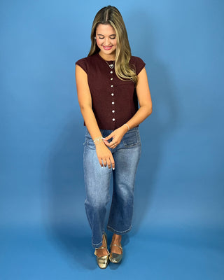 A woman modeling a sleeveless burgundy knit top with a button-down front, paired with blue jeans and metallic gold flats, standing against a solid blue studio backdrop.