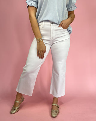 Person wearing a light blue striped shirt, white pants, and gold sandals on a pink background