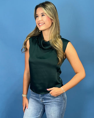 A woman with blonde hair wearing a dark green sleeveless cowl-neck top and light blue jean