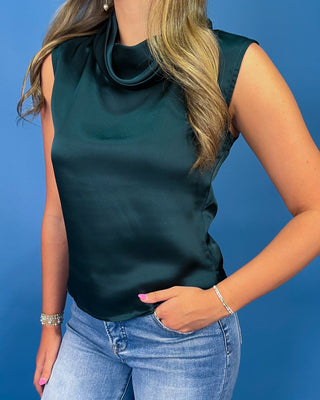 A woman wearing a dark green satin sleeveless top with a cowl neck, paired with light blue jean