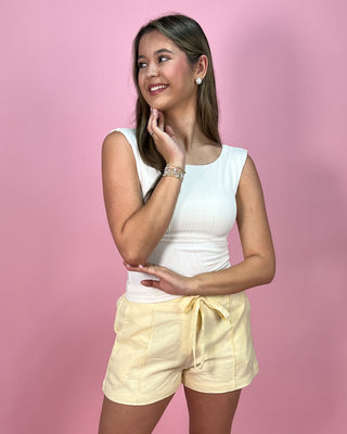 Woman wearing a white sleeveless top and yellow shorts against a pink background