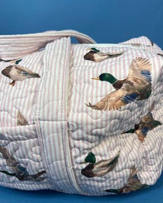 Ida Mae Home quilted duck print duffle bag with striped fabric, padded handles, spacious zip-top compartment, available in pink or blue.