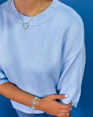 A close-up of a person wearing a light blue knit crewneck sweater, styled with a gold heart necklace and stacked bracelets against a solid blue background.