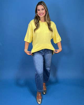 A woman modeling a bright yellow short-sleeve knit top with a ruffled hem, paired with blue jeans and metallic shoes, standing against a solid blue studio backdrop.