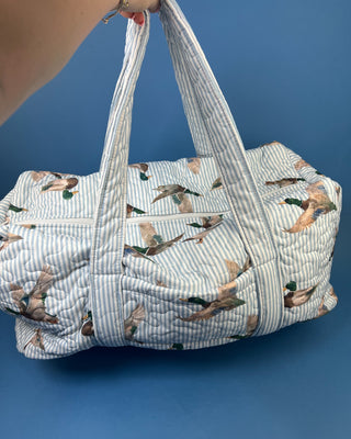Ida Mae Home quilted duck print duffle bag with striped fabric, padded handles, spacious zip-top compartment, available in pink or blue.