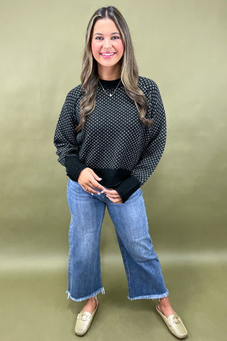 Woman wearing a black patterned top and blue jeans against a green background