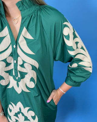 A close-up of a woman wearing a teal green satin-like blouse with large cream-colored baroque-style embroidery on the chest and sleeves