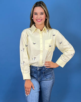 A woman modeling a pale yellow long-sleeve button-up shirt with small Mallard duck prints, paired with blue jeans and metallic shoes, standing against a solid blue studio backdrop.