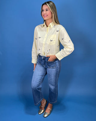 A woman modeling a pale yellow long-sleeve button-up shirt with small Mallard duck prints, paired with blue jeans and metallic shoes, standing against a solid blue studio backdrop.
