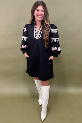 Woman wearing a black dress with white embroidery and white knee-high boots on a green background
