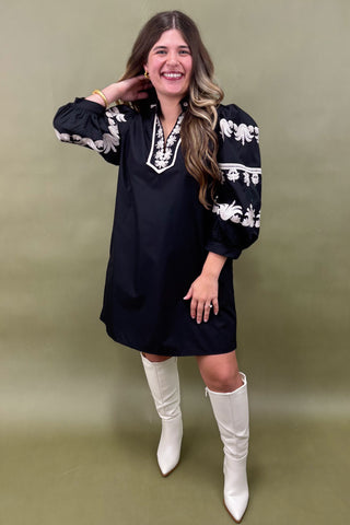 Woman wearing a black dress with white patterns and white boots against a green background