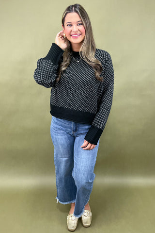 Woman wearing a black patterned sweater and blue jeans against a beige background
