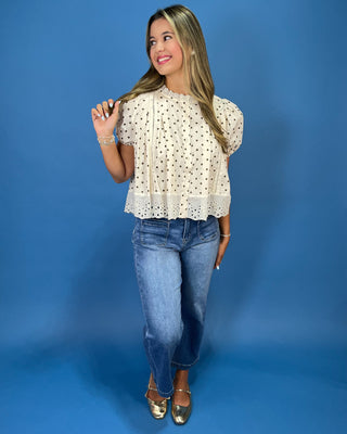 A woman modeling a cream eyelet short-sleeve blouse with black polka dots, paired with blue jeans and metallic gold flats, standing against a solid blue studio backdrop.