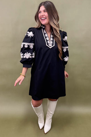 Woman wearing a black dress with white embroidery and white boots on a green background