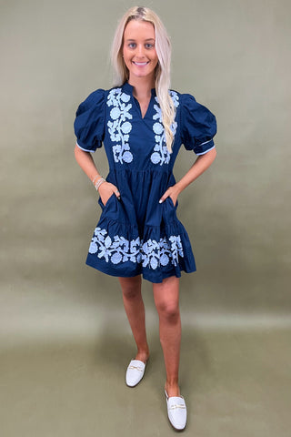 Woman wearing a navy dress with white floral embroidery against a beige background