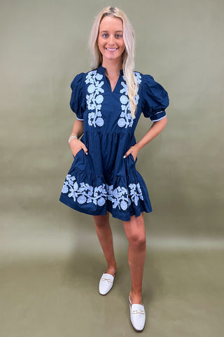 Woman wearing a blue dress with white floral embroidery against a plain background
