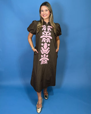 A woman modeling a brown midi dress with puff sleeves and a pink damask pattern down the front