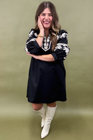 Woman wearing a black dress with white patterns and white boots against a green background
