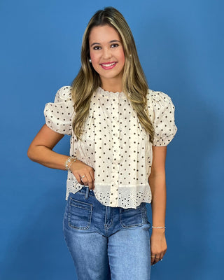 A woman modeling a cream eyelet blouse with black polka dots and puff sleeves, paired with blue jeans, against a solid blue studio background.