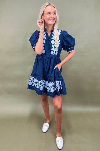 Woman wearing a blue dress with white floral embroidery against a beige background
