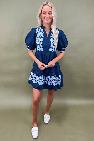 Woman wearing a navy dress with white floral embroidery against a plain background