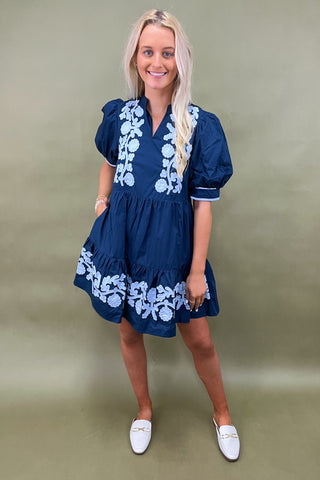 Woman wearing a navy dress with white floral embroidery against a beige background