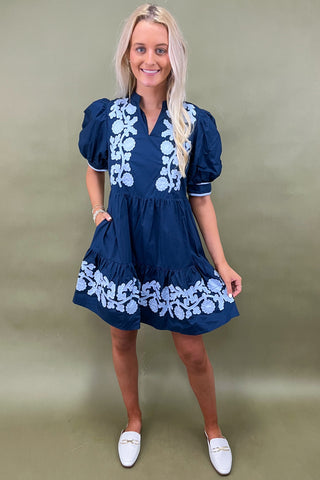 Woman wearing a navy dress with white floral embroidery against a green background