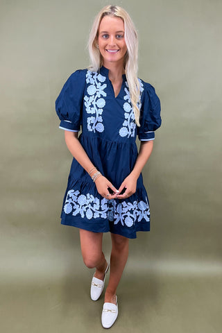 Woman wearing a blue dress with white floral embroidery against a green background