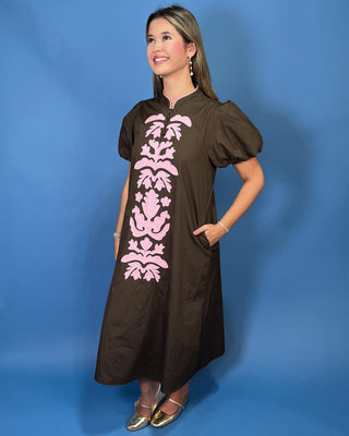 A woman modeling a brown midi dress with puff sleeves, a mock neck, and a pink damask pattern down the front