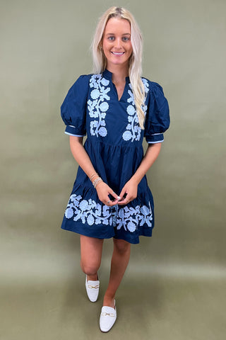 Woman wearing a blue dress with white floral embroidery against a green background
