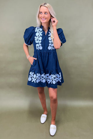 Woman wearing a blue dress with white floral embroidery against a plain background