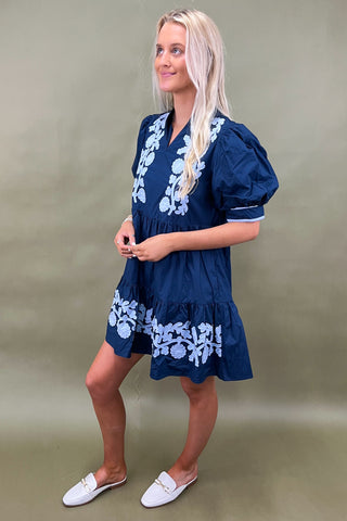 Woman wearing a navy dress with white floral embroidery against a green background