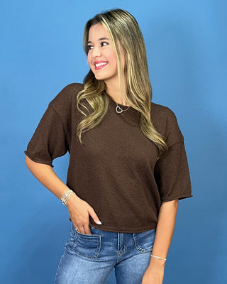 A woman wearing a brown short-sleeve knit top and blue jeans, standing against a solid blue studio background.