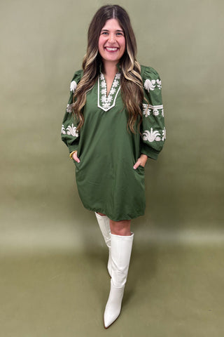 Woman wearing a green dress with white embroidery and white boots on a green background