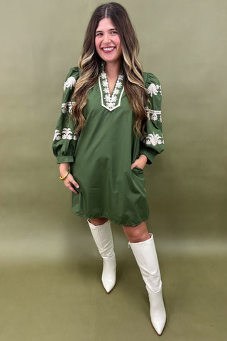 Woman wearing a green dress with white embroidery and white knee-high boots on an olive green background