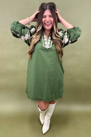 Woman wearing a green dress with floral sleeves and white boots against a green background