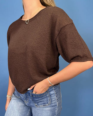 A woman wearing a brown short-sleeve knit sweater and blue jeans, standing against a solid blue background.