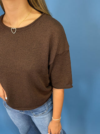 A woman wearing a brown short-sleeve knit top with a wide scoop neckline, paired with light blue jeans, standing against a solid blue background.