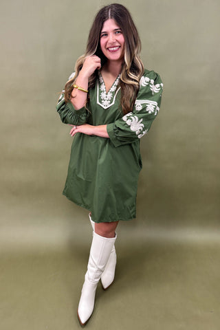 Woman wearing a green dress with white floral patterns and white boots against an olive green background