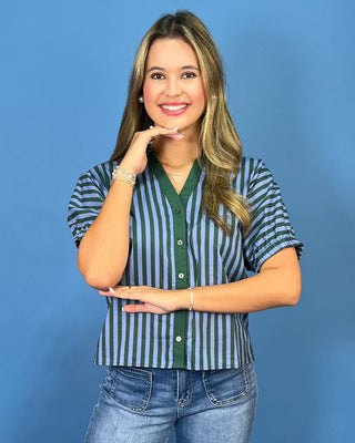 A woman modeling a short-sleeve button-up shirt with vertical green and blue stripes, paired with blue jeans, set against a solid blue studio background.
