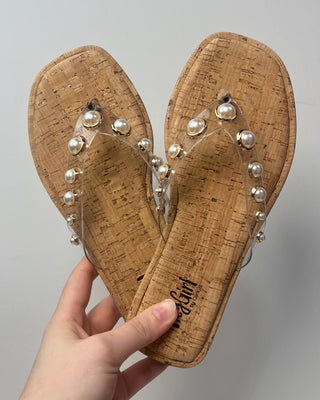 Pair of cork sandals with pearl embellishments held by a hand against a gray background