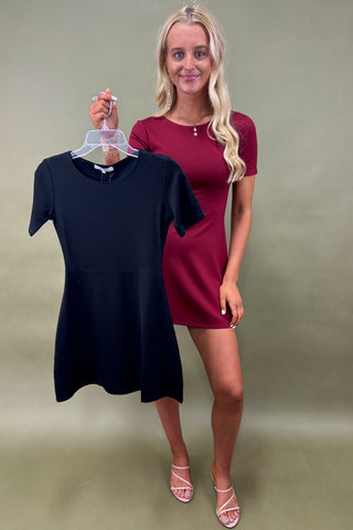 Woman holding a black dress on a hanger next to a maroon dress against a plain background