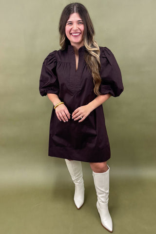 Woman wearing a dark purple dress with puffed sleeves and white knee-high boots against a green background