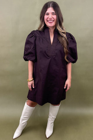 Woman wearing a dark purple dress with puff sleeves and white boots against an olive green background