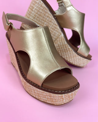 Gold wedge sandal with straw sole held by a hand against a pink background