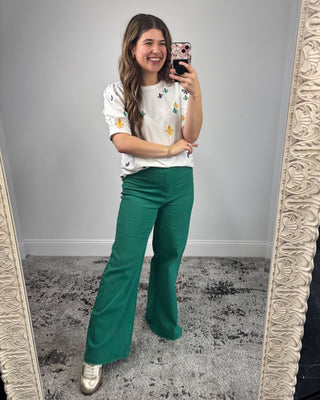 Woman taking a mirror selfie wearing a white floral top and green pants.
