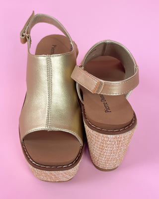 Gold wedge sandal with straw sole held by a hand against a pink background