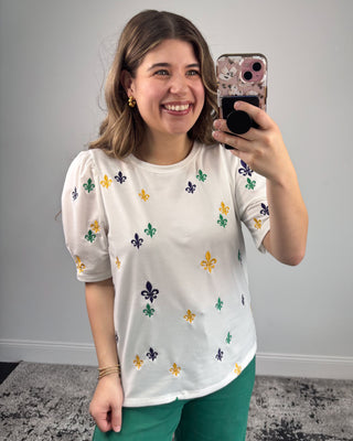Woman taking a mirror selfie wearing a white shirt with colorful patterns and green pants.